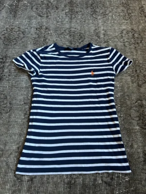 Randig t-shirt från Ralph Lauren Sport - Säljer en snygg marinblå och vit randig t-shirt från Ralph Lauren Sport. T-shirten har korta ärmar, rund halsringning och den klassiska lilla loggan broderad på bröstet. Tillverkad i 100% bomull för skön känsla.