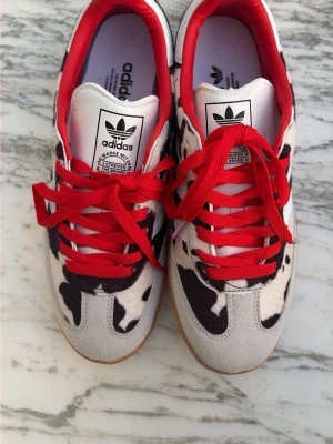 Adidas Samba OG sneakers med koskinnsmönster - Unika Adidas Samba OG  sneakers med svartvitt koskinnsmönster, röda skosnören och röd insida. Klassisk Adidas-logga på plösen och vita detaljer på sidan. Yttermaterial i mocka och textil, sula i gummi. Mina blå jeans har gnuggat av lite färg på mockan när jag provat dem. Går att ta bort. Säljer billigt så jag kan köpa nya skor snabbt för dessa var för stora för mig. True to size. 
