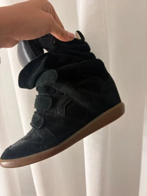 Svarta Isabel Marant sneakers - Säljer ett par svarta höga sneakers från Isabel Marant med cool design och kardborreband. Skorna har en brun gummisula och är tillverkade i mocka och textil. Perfekta för dig som vill ha en unik och trendig look. Köpta nya för 6000 i butik. Vid snabbt köp kan jag sänka priset  