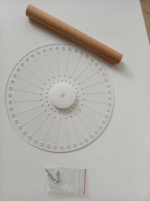 Tårtbottendelare i trä och plast - .Detta makraméverktyg är utformat för att skapa runda makraméprojekt. Verktyget består av en rund platta med numrerade hål längs kanten, vilket underlättar jämn fördelning av trådar. I mitten finns en skumkärna. En träpinne och skruvar medföljer.