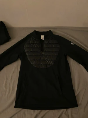 Svart tröja med quiltad front från Nike - Svart långärmad tröja från Nike med quiltad, vadderad frontpanel och half zip-dragkedja. Tröjan har en sportig look och är perfekt för kyligare dagar. Nike-logga på ärmen och hög krage för extra komfort.