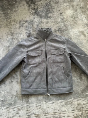 Grå mockaliknande jacka med fickor - Snygg grå jacka i mockaliknande material med hög krage och dragkedja framtill. Två bröstfickor med lock och två sidofickor ger en clean och modern look. Perfekt för dig som gillar minimalistisk stil och vill ha något som sticker ut lite extra.