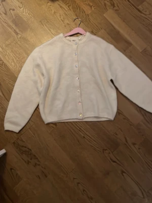 Beige kofta med pärlemorknappar - Mysig beige kofta med rund halsringning och raka ärmar. Koftan har pärlemorskimrande knappar framtill och en enkel, stilren design. Perfekt att slänga över en topp för en avslappnad look.