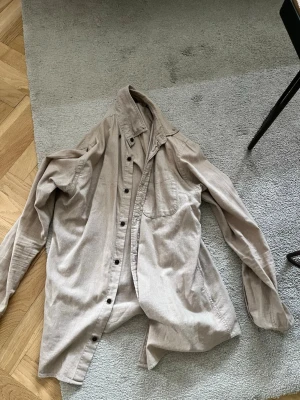 Beige manchesterskjorta med knappar - Säljer en beige skjorta i mjuk manchester med klassisk krage och knappar framtill. Skjortan har en bröstficka och långa ärmar. Perfekt för dig som gillar en avslappnad och stilren look.