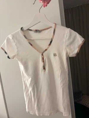 Vintage Burberry t shirt - Äkta men lite gammal/sliten. Vintage 2000’s Burberry  t-shirt med korta ärmar och v-ringning. T-shirten har rutiga detaljer längs halsringningen och ärmsluten samt knappar framtill. Liten broderad logga på bröstet. Tillverkad i mjuk bomull och har en normal passform.