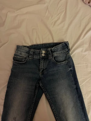 Mörkblå bootcut jeans med dubbla knappar - Säljer ett par mörkblå bootcut jeans med snygg tvätt och dubbla knappar i midjan. Jeansen har klassiska fem fickor och subtila slitningar framtill. Perfekta för dig som gillar en lite vidare passform nedtill och vill ha en tidlös look.