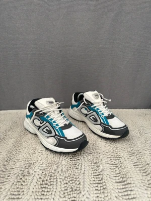 Vita och blå sneakers med svarta detaljer - Säljer ett par snygga sneakers i vitt med blå och svarta detaljer. Skorna har en sportig design med mesh- och syntetmaterial, vita snören och en rejäl sula. Perfekta för dig som gillar streetstyle och vill ha något som sticker ut.