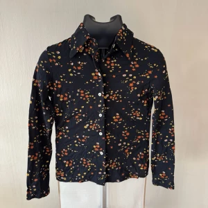 Phard Svart skjorta med blommigt mönster - Snygg svart skjorta med små blommor i orange, gul och vit. Skjortan har långa ärmar, klassisk krage och knäppning framtill med vita knappar. Perfekt för dig som vill ha en färgglad touch i garderoben.0369