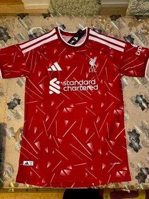Liverpool home röd matchtröja 26/27 Adidas player version - Säljer en röd Liverpool matchtröja från Adidas med vita detaljer och grafiskt mönster. Tröjan har klubbmärke, sponsorlogga och nummer 7 med namnet Wirtz på ryggen. Perfekt för fotbollsfans som vill sticka ut på planen eller läktaren. Home 26/27 player version 