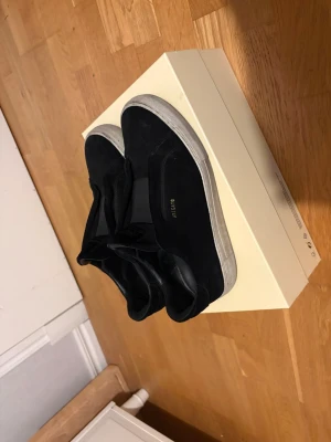 Svarta Axel Arigato sneakers - Säljer ett par svarta sneakers från Axel Arigato med vit sula och diskret logga i guld på sidan. Skorna har en stilren och minimalistisk design i mocka och läder, perfekta för dig som gillar en clean look.