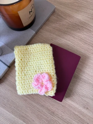Gul virkad passfodral med blomma✨ - Handgjort virkat passfodral i mjukt gult garn med en söt rosa blomma på framsidan🌸Perfekt för att skydda ditt pass och ge det en personlig touch. Fodralet är flexibelt och har en enkel design som gör det lätt att använda✨