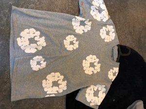 Grå t-shirt med vita blommor - Säljer en grå t-shirt från Denim Tears i storlek M. T-shirten har korta ärmar och är dekorerad med stora vita blommor över hela plagget. Perfekt för dig som vill sticka ut med ett unikt mönster och skön passform.
