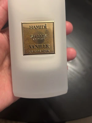 Hamidi Shams Vanille parfym - En elegant parfym från Hamidi, Shams Edition Vanille L'Eau De Aqua. Flaskan är frostad vit med en lyxig guldfärgad etikett framtill. Doften har tydliga inslag av vanilj och flaskan har en modern, stilren design som känns exklusiv.