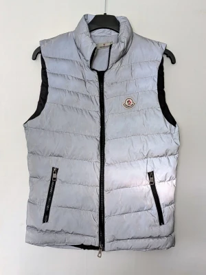 Silvrig  dunväst från Moncler - Snygg silvrig dunväst från Moncler med hög krage och två dragkedjefickor framtill. Västen har quiltad design och klassisk Moncler-logga på bröstet. Perfekt för lager-på-lager och enkel att matcha till olika outfits.