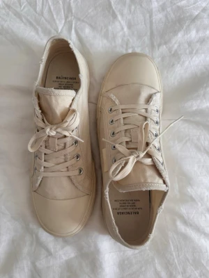 Beige sneakers från Balenciaga - Säljer ett par beige sneakers från Balenciaga med slitna kanter och klassisk låg design. Skorna har vita snören, silverfärgade öljetter och är tillverkade i canvas med gummisula. Perfekta för en avslappnad och trendig stil.