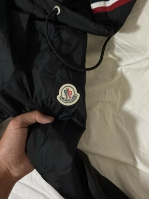 Moncler windbreaker NFC - Snygg svart vindjacka från Moncler med dragkedja, två framfickor med knappar och justerbar huva med snörning. Jackan har Moncler-logga på ärmen och vita/röda detaljer på huvan. Perfekt för blåsiga dagar och streetwear-stil.