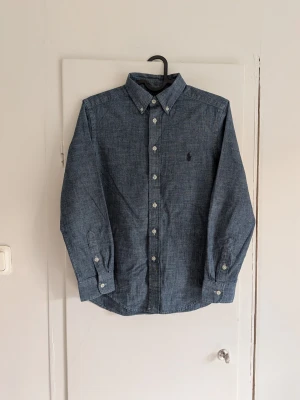 Blå skjorta från Polo Ralph Lauren - Snygg blå skjorta från Polo Ralph Lauren med klassisk button-down krage och broderad logga på bröstet. Skjortan har långa ärmar och knappar framtill. Perfekt till jeans eller chinos för en clean look. Skjortan har en fläck (se sista bilden).