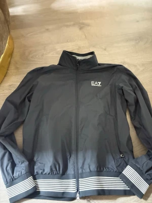 Svart vindjacka från EA7 Emporio Armani - Snygg svart vindjacka från EA7 Emporio Armani med hel dragkedja, ståkrage och vita ränder på muddar och nederkant. Jackan har två sidofickor och EA7-logga på bröstet. Tillverkad i lätt och smidigt material som passar perfekt till streetwear. ÄKTA!