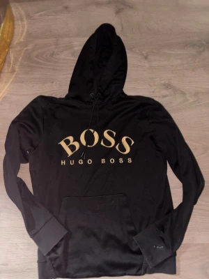 Svart hoodie från Hugo Boss - Snygg svart hoodie från Hugo Boss med stor guldfärgad BOSS-logga på bröstet. Klassisk huva med dragsko och stor magficka framtill. Tillverkad i mjukt material och har långa ärmar med muddar. Perfekt för en avslappnad och trendig look.