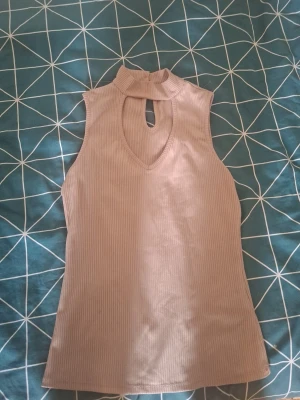 Beige ribbad ärmlös topp med hög krage - Säljer en beige ribbad topp med hög krage och snygg keyhole-detalj både fram och bak. Toppen är ärmlös och har en slim passform som sitter snyggt på kroppen. Perfekt att styla med jeans eller kjol för en trendig look.