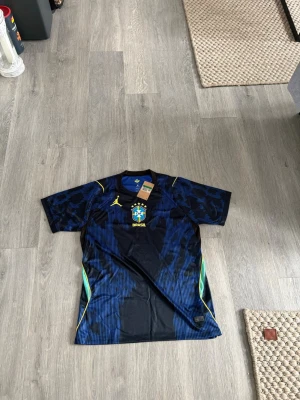Blå Brasilien matchtröja - Snygg blå matchtröja för Brasilien med gul logga och emblem på bröstet. Tröjan har korta ärmar, svarta och blå mönster samt gula och gröna detaljer på sidorna. Perfekt för fotbollsträning eller att bära till vardags.