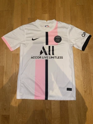 PSG matchtröja vit rosa svart Oanvänd  - Snygg PSG matchtröja i vitt med rosa och svart vertikal rand, svart Nike-logga och PSG-emblem på bröstet. Korta ärmar med rosa och svart detalj på ärmsluten. Tillverkad i lätt och ventilerande material, perfekt för fotbollsträning eller sport.