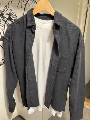 Mörkgrå manchester overshirt - Oanvända, Snygg mörkgrå overshirt i manchester med klassisk krage och bröstficka. Skjortan har långa ärmar och knäppning framtill. Perfekt att bära öppen över en t-shirt för en avslappnad look.