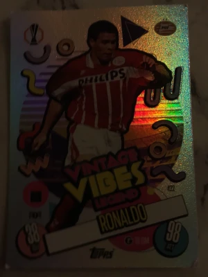 Vintage Vibes Legend Ronaldo kort - Säljer ett glittrigt samlarkort från Topps med fotbollsspelaren Ronaldo i PSV Eindhovens rödvita matchtröja. Kortet har färgglada retrodetaljer och texten 'Vintage Vibes Legend'. Perfekt för dig som gillar fotboll och samlarkort.