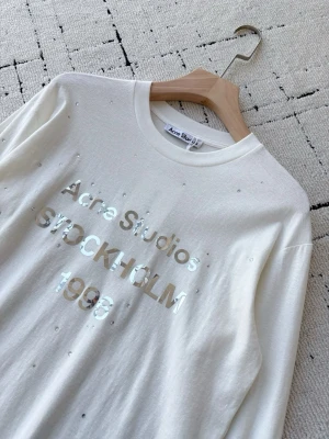 Vit Acne Studios t-shirt med tryck - Snygg vit t-shirt från Acne Studios med glansigt tryck framtill där det står 'Acne Studios STOCKHOLM 1996'. T-shirten har små dekorativa stenar över hela plagget och är gjord i mjukt material. Perfekt för dig som gillar minimalistisk men ändå unik stil.