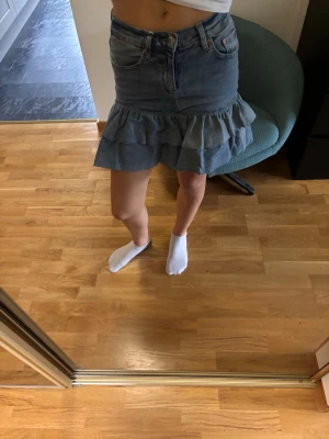 Blå jeanskjol med volang - Säljer en blå jeanskjol med volangkant nedtill. Kjolen har klassiska jeansfickor fram och bak, samt knapp och dragkedja i midjan. Är ett par jeans som jag har sytt om till en kjol, nu kommer den aldrig till användning, inte använda mycket!
