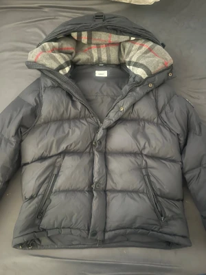 Svart dunjacka från Burberry  - Säljer en svart dunjacka från Burberry med avtagbara armar som kan även bli en väst  och klassiskt rutigt foder i grått, rött och vitt. Jackan har två dragkedjeförsedda fickor, tryckknappar och Burberry London England-logga på ärmen. Säljs pga storlek.