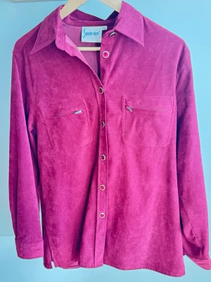 Vintage skjorta vinröd strl M - Snygg rosa skjorta i manchester från Souerdame. Skjortan har långa ärmar, klassisk krage, knappar framtill och två bröstfickor med dragkedja. Perfekt för dig som vill sticka ut med färg och mjukt material Vintage Skjorta med långa ärmar och krage. Försluts med knappar framtill. Två bröstfickor med dragkedja. Ärmsluten har knappar. Skjortan har slitsar i sidorna. Bröstvidd: ca 49 cm Längd 72 cm ca  Superfin skick utan några skavanker. Längre modell