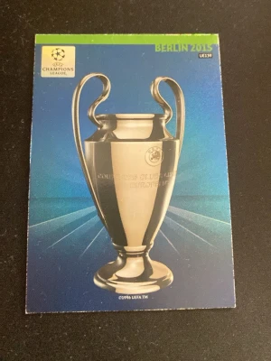 UEFA Champions League klistermärke - Klistermärke med motiv av Champions League-pokalen från Berlin 2015. Bakgrunden är blå med grönt fält upptill och texten 'BERLIN 2015'. Perfekt för samlare av fotbollsrelaterade böcker eller album.