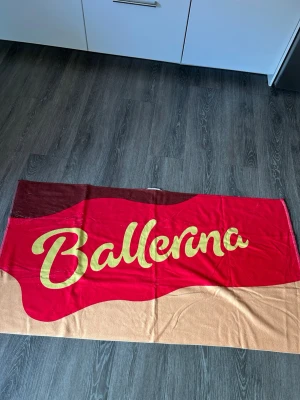 Ballerina strandhandduk med tryck - Säljer en stor strandhandduk med Ballerina-tryck i rött, beige och brunt. Handduken har en mjuk frottésida i vitt på baksidan och färgglad framsida med texten 'Ballerina' i gult. Perfekt för stranden eller poolen. Helt ny!!