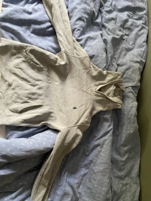 Ljusgrå hoodie med huva - Ljusgrå hoodie med klassisk känguruficka och huva. Tröjan har en liten broderad logga på bröstet och är tillverkad i mjukt bomullsmaterial. Perfekt för en avslappnad och stilren look.