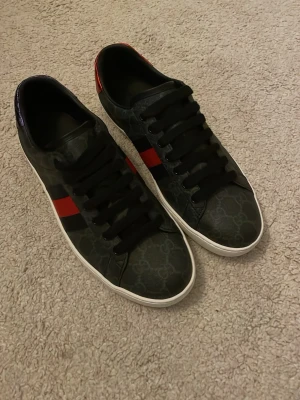 Svarta Gucci sneakers med röd rand - Säljer ett par svarta Gucci sneakers med ikoniskt GG-mönster och en röd och marinblå rand på sidan. Skorna har svarta skosnören, vit sula och är tillverkade i skinn. Perfekta för dig som vill ha en stilren och lyxig look.