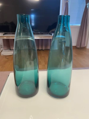 Två blågröna handgjorda glasvaser - Säljer två stilrena blågröna glasvaser med handgjort kvalitetsmärke. Vaserna har en avlång, mjukt böjd form och passar perfekt som dekoration eller till snittblommor. Färgen är transparent blågrön och glaset har en tjock botten.