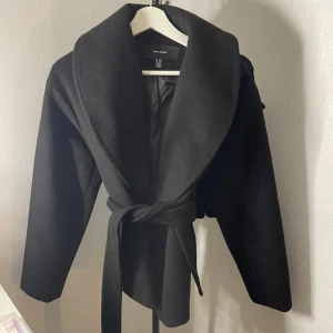 Svart kappa från Vero Moda XS - Stilren svart kappa från Vero Moda med bred sjalkrage och knytskärp i midjan. Kappan har en klassisk, rak passform och är helfodrad för extra komfort. Perfekt för kyliga dagar och enkel att matcha med olika outfits.