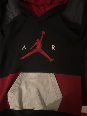Svart och vinröd hoodie med AIR-tryck - Säljer en svart och vinröd hoodie med stor AIR-logga och ikoniskt baskettryck både fram och bak. Hoodien har en stor magficka i grått och en rymlig huva. Perfekt för dig som gillar streetwear och sportig stil.