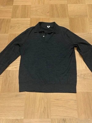 Mörkgrå stickad pikétröja - En timeless mörkgrå stickad pikétröja med lång ärm och klassisk krage. Tröjan har två knappar vid halsen och ribbade muddar vid ärmslut och nederkant. Perfekt för en avslappnad men ändå uppklädd look.