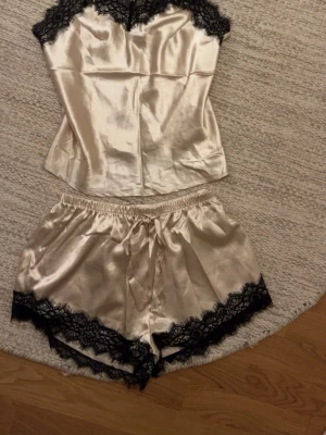 Silkeslent pyjamasset med spetsdetaljer - Snyggt pyjamasset i ljus champagnefärgad satin med svarta spetsdetaljer. Setet består av ett linne med tunna axelband och matchande shorts med elastisk midja. Både linne och shorts har dekorativ svart spets längs kanterna för en lyxig känsla.