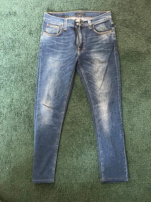 Blå Nudie Jeans med tvätt - Säljer ett par blå jeans från Nudie med snygga slitningar och tvättade detaljer framtill. Jeansen har klassisk femficksmodell, smal passform och kontrastsömmar. Bakfickorna har dekorativa sömmar. Perfekta till en avslappnad stil.