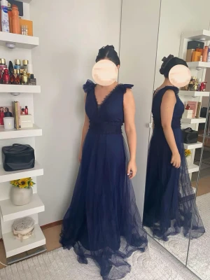 Marinblå lång klänning med volang - Elegant marinblå långklänning med v-ringning och volangdetaljer vid axlar och urringning. Klänningen har markerad midja och är tillverkad i ett tunt, svepande material som ger en lyxig känsla.