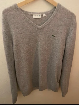Grå v-ringad tröja från Lacoste - Mjuk och stilren grå stickad tröja från Lacoste med v-ringning och den klassiska gröna krokodilloggan på bröstet. Tröjan har långa ärmar och ribbade muddar vid ärmslut och nederkant. Perfekt för lager på lager och enkel att matcha.