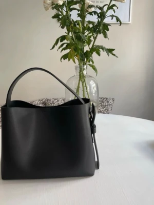Svart stilren handväska i skinnimitation - Snygg svart handväska med minimalistisk design och bred rem. Väskan har en boxig form och är tillverkad i slät skinnimitation. Perfekt för dig som gillar enkel och modern stil. Spänndetalj på sidan ger en extra touch.