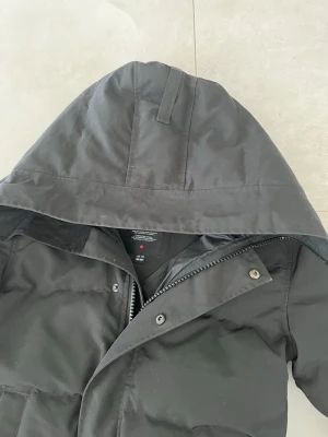 Svart dunjacka med huva Canada Goose - Säljer en svart dunjacka från Canada Goose med stor huva och dragkedja samt knappar framtill. Jackan har en klassisk patch på ärmen och två stora fickor framtill. Perfekt för kalla vinterdagar och riktigt varm tack vare dunfyllningen.