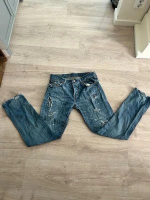 Blå slitna jeans med raka ben - Säljer ett par blå jeans med slitningar och hål på flera ställen. Jeansen har raka ben, klassisk femficksmodell och knappgylf. Materialet är denim i bomull och de har en avslappnad, vintage look. Perfekta för dig som gillar en edgy och avslappnad stil.