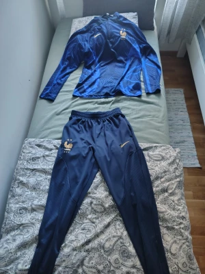Blått Frankrike träningsset Nike - Säljer ett blått träningsset från Nike med Frankrikes landslagslogga. Setet består av en långärmad tröja och matchande byxor i funktionsmaterial. Båda plaggen har gulddetaljer och är perfekta för fotbollsträning eller gymmet.