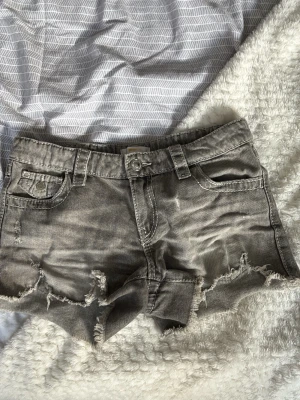 Grå slitna jeansshorts med detaljer på bakfickorna - Säljer ett par grå jeansshorts med slitna kanter och råa detaljer. Shortsen har fem fickor, bälteshällor och snyggt broderade bakfickor med ljusblå och vita detaljer. Perfekta för sommaren och ger en avslappnad vibe.