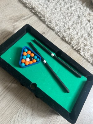 Mini biljardbord med tillbehör - Säljer ett mini biljardbord i svart och grönt med två svarta köer, en blå triangel och färgglada bollar i orange, blått, vitt och svart. Perfekt för spel hemma med kompisar eller familj. Bordet är kompakt och lätt att ställa undan. 